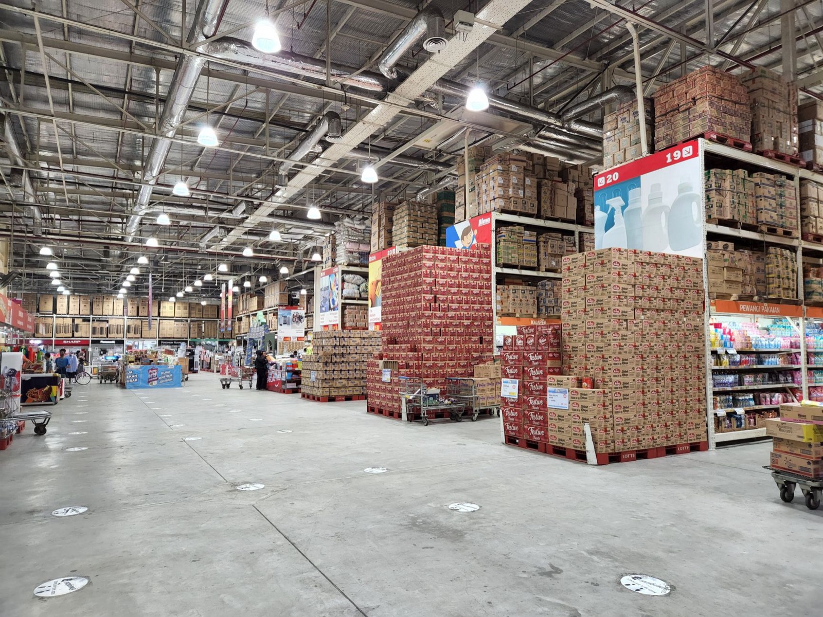 A large wholesale warehouse interior filled with stacked merchandise and shoppers browsing the aisles.